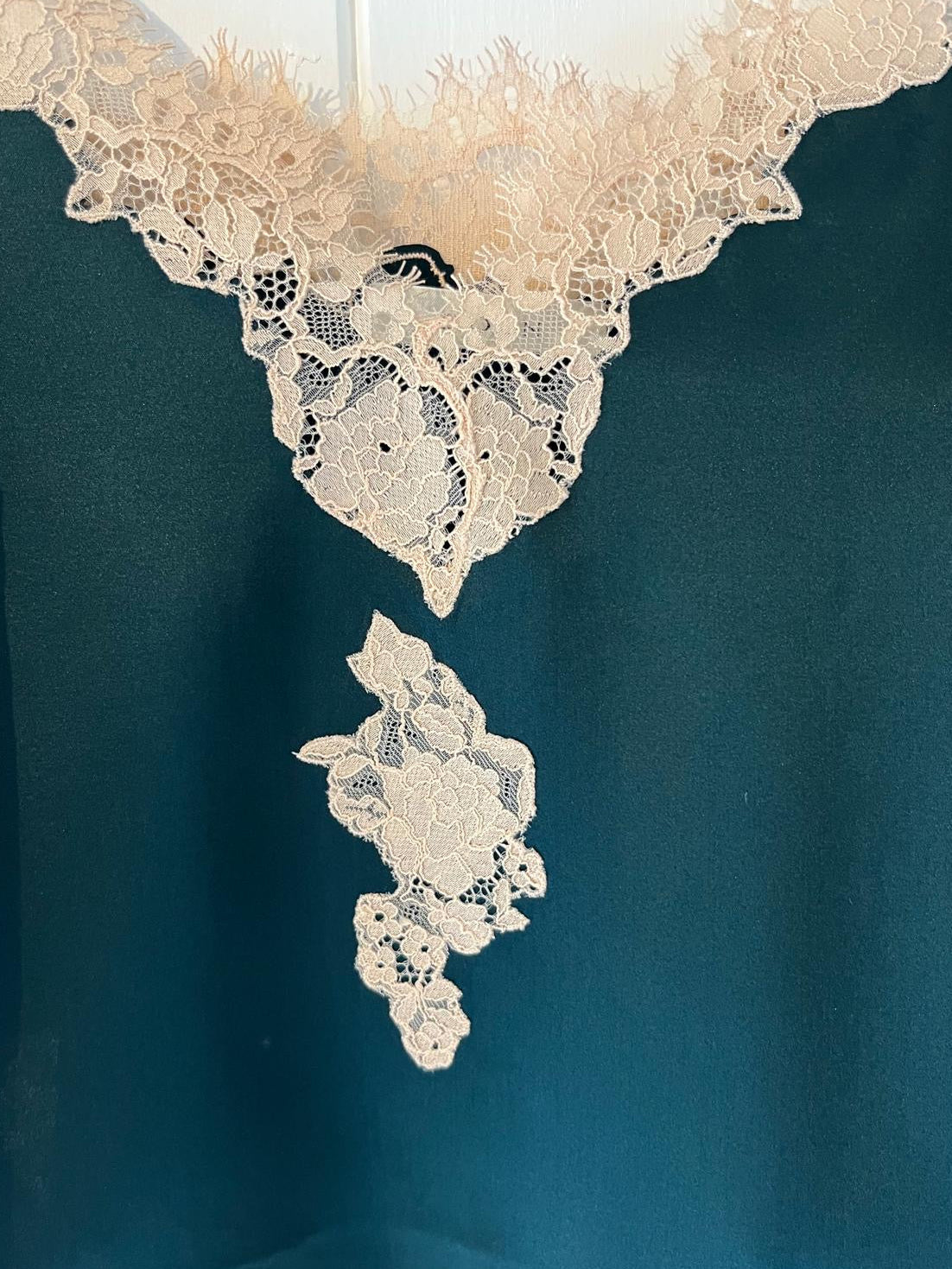Close-up of a dark teal garment featuring intricate cream lace applique detailing along the neckline and a matching lace motif below it.