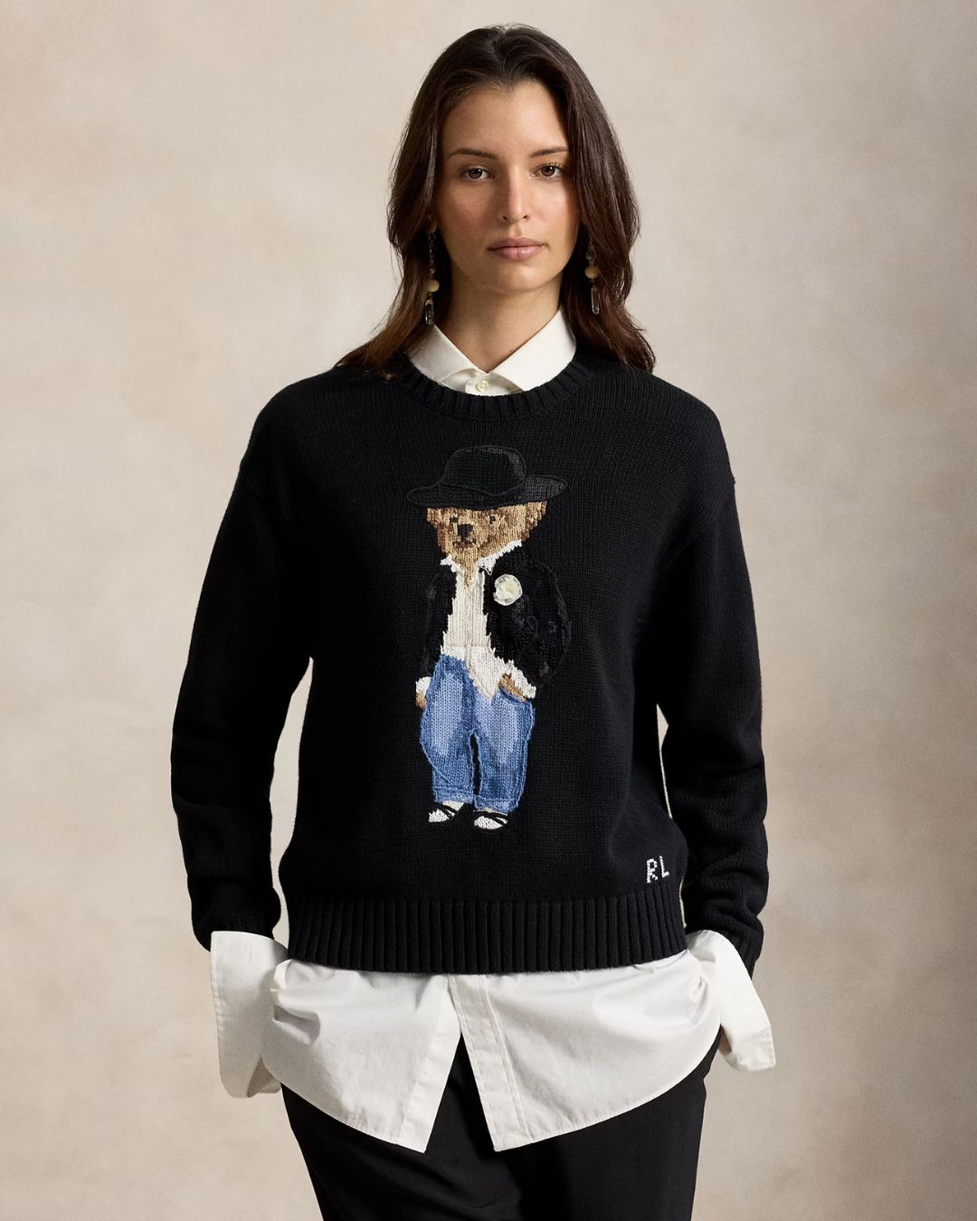 A woman wearing a black knit sweater featuring a large embroidered teddy bear in a hat and blue jeans on the front, styled over a white collared shirt and black pants against a neutral beige background.