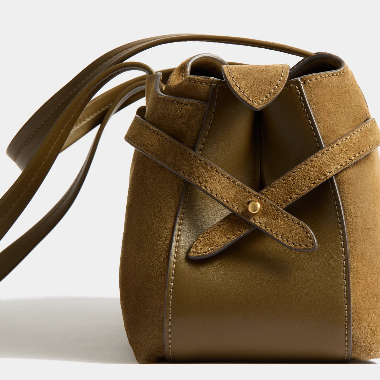 A close-up of a brown leather and suede bucket bag with a drawstring closure and a gold-tone snap button, set against a clean white background.