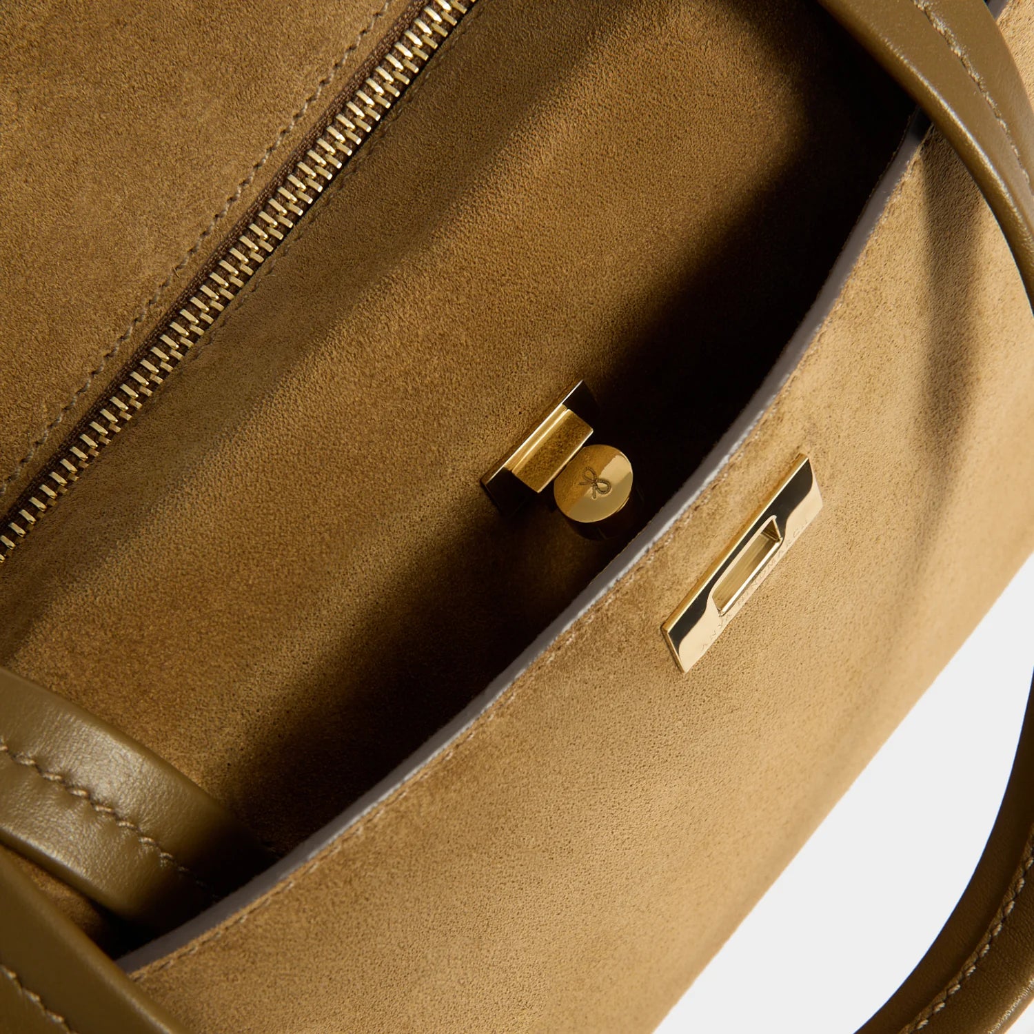Close-up of the interior of a structured handbag in warm tan suede or leather, showing a gold-tone zipper, turn-lock hardware, and a metal logo plate on the flap against a white background.