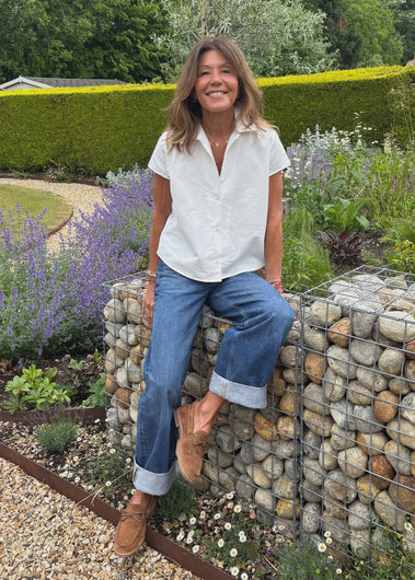 Woman in a white shirt and blue jeans sitting on a stone wall in a garden.