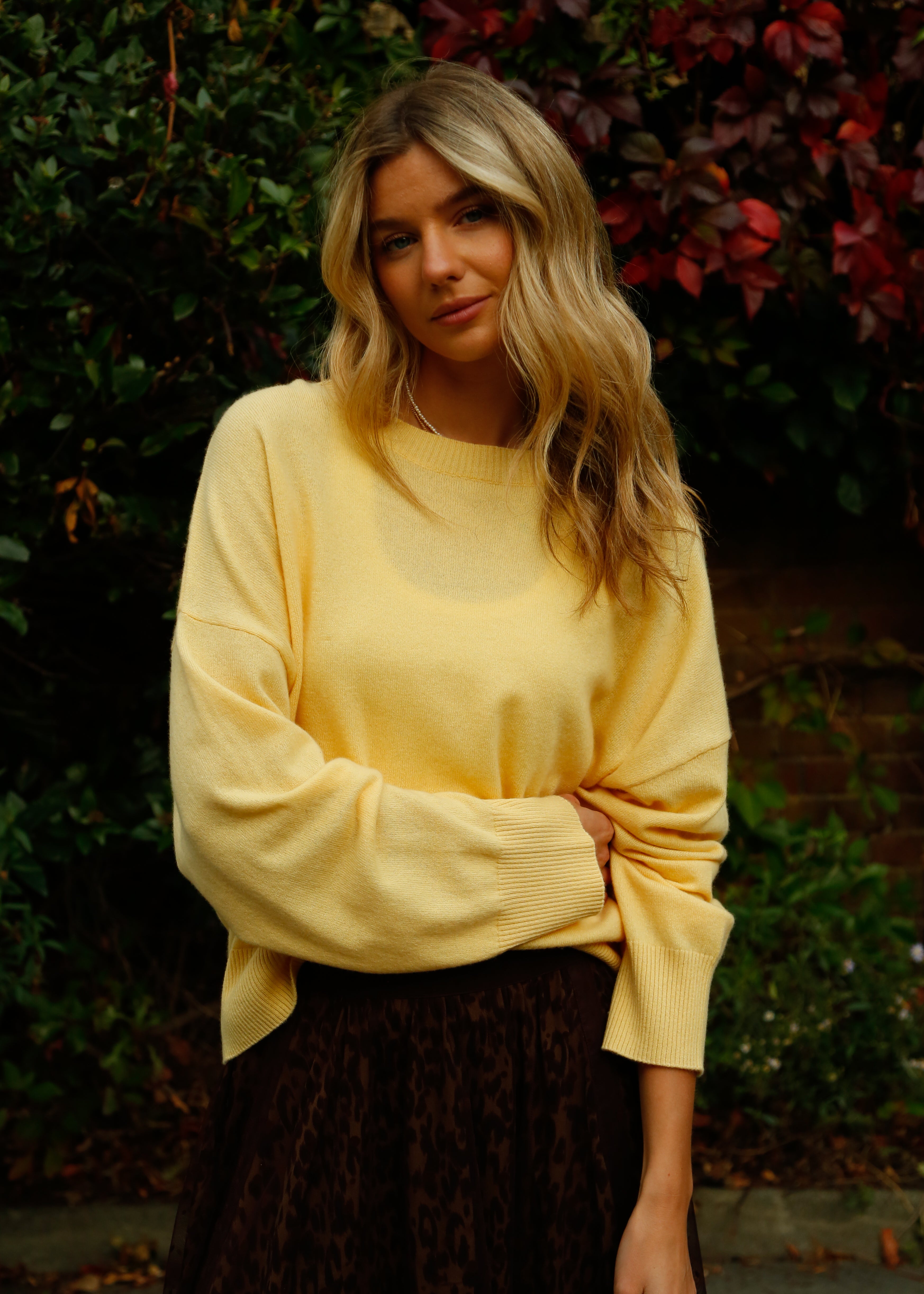 Woman wearing a yellow sweater standing in front of green foliage
