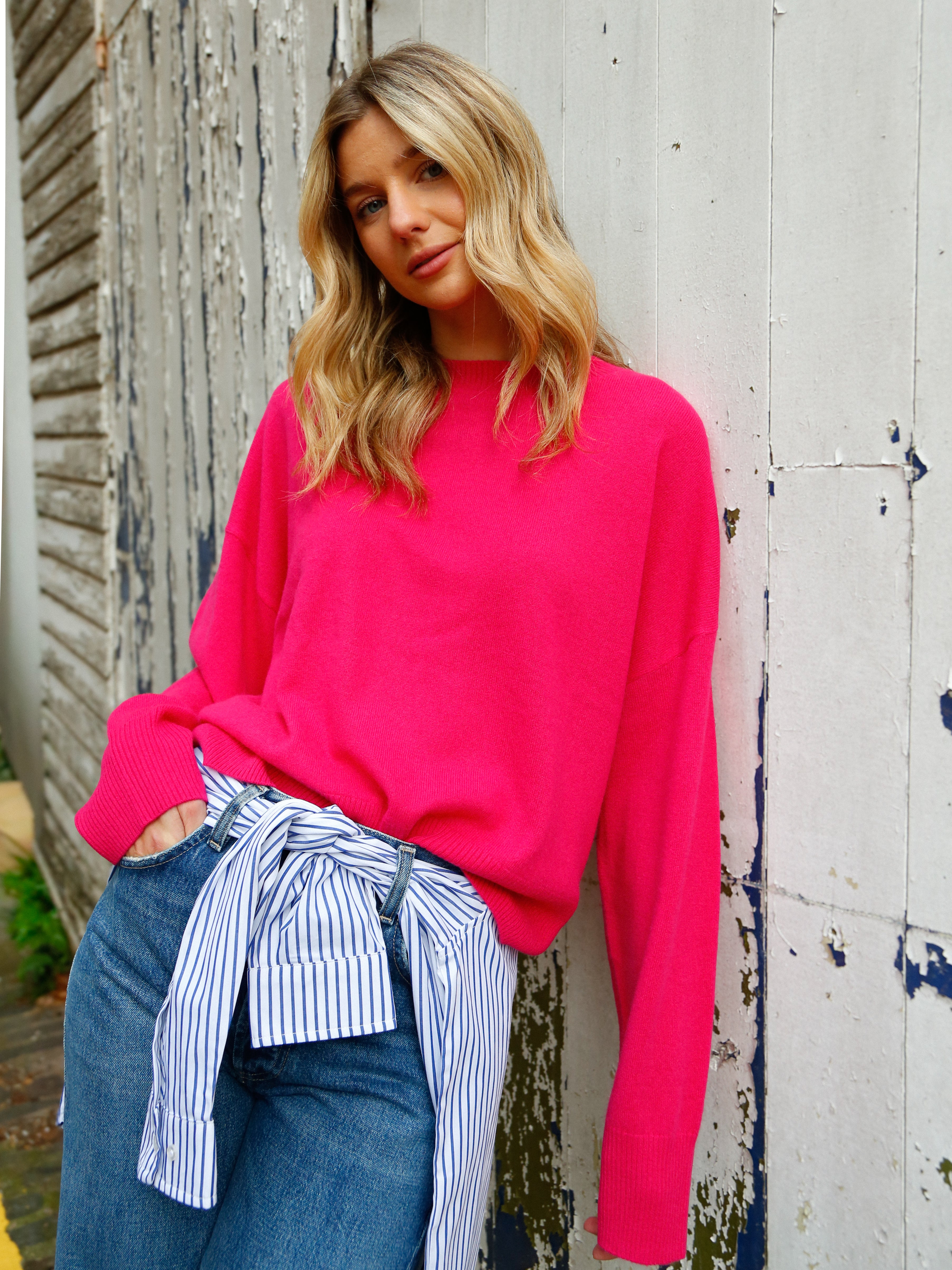 Woman wearing a bright pink sweater with a striped shirt tied around her waist, standing against a rustic wooden wall.