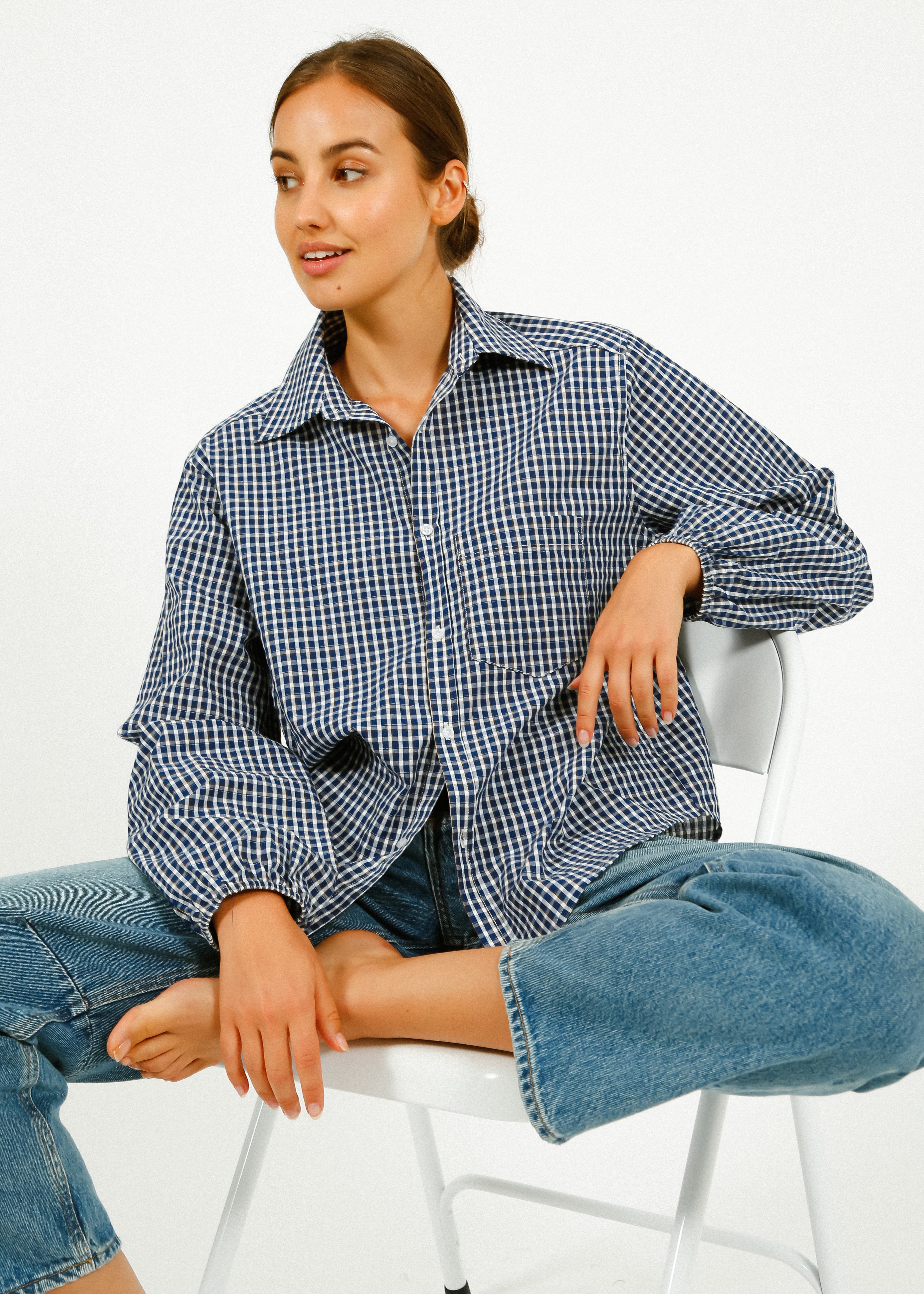 Woman wearing a blue checkered shirt and jeans sitting on a white chair.