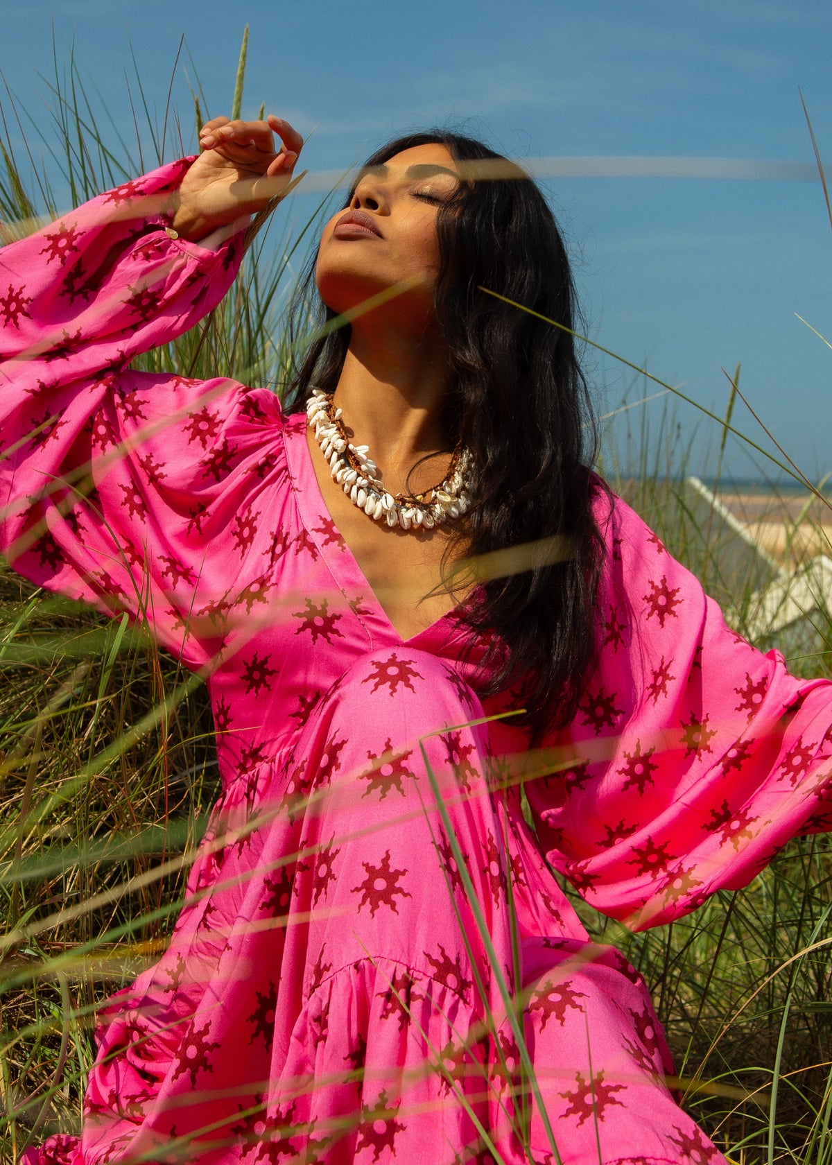 A woman sitting in a field of tall grass wearing a pink sleeveless maxi dress with a gathered blouson sleeve and a fitted bust, posing with one hand raised to her face.