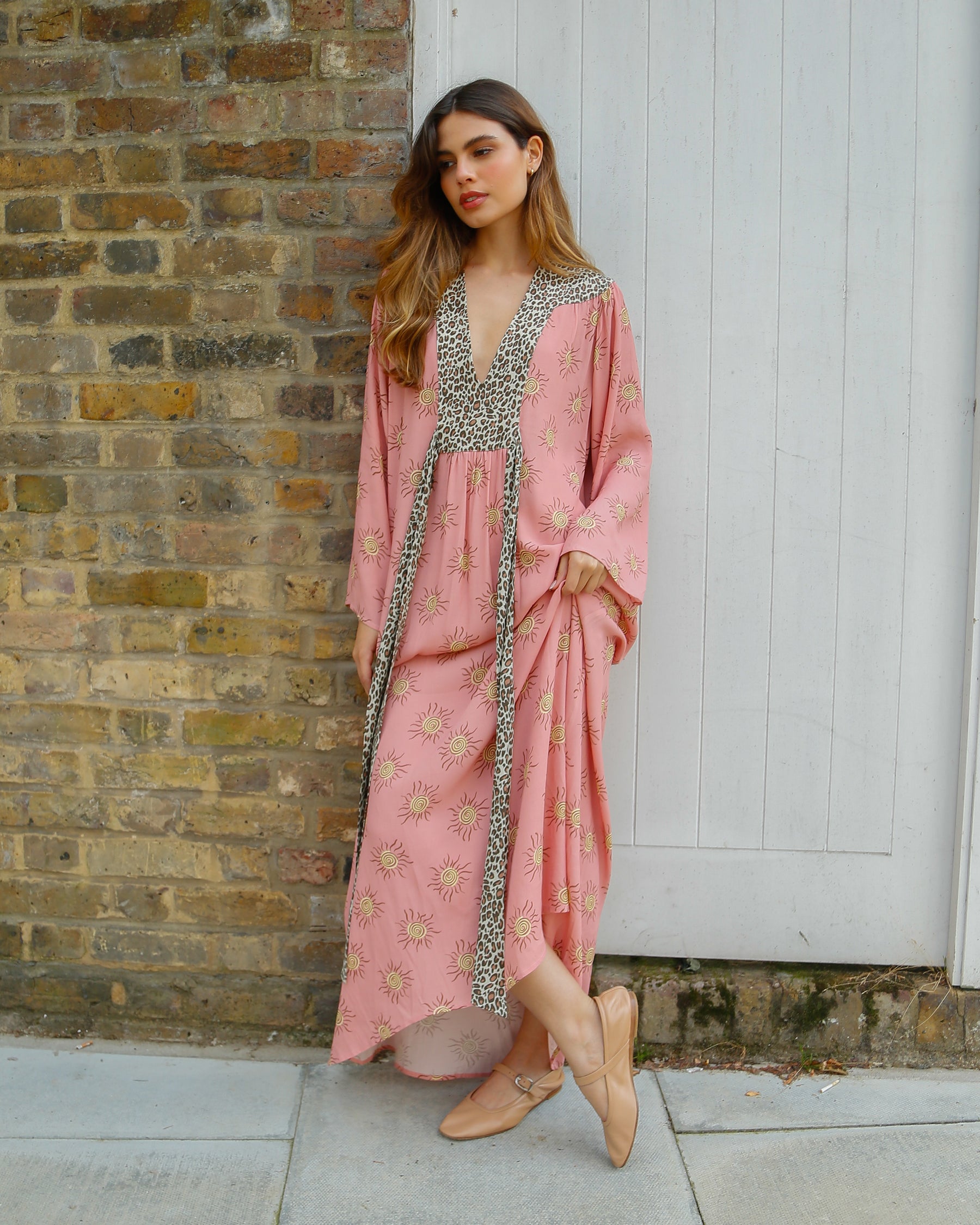 A woman standing by a brick wall and white door wearing a long pink maxi dress with leopard print detailing.