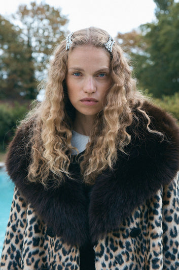 A model stands on a stone pathway in a formal garden wearing a leopard-print coat with a large dark fur collar and cuffs, a black dress, white tights, and burgundy heels.
