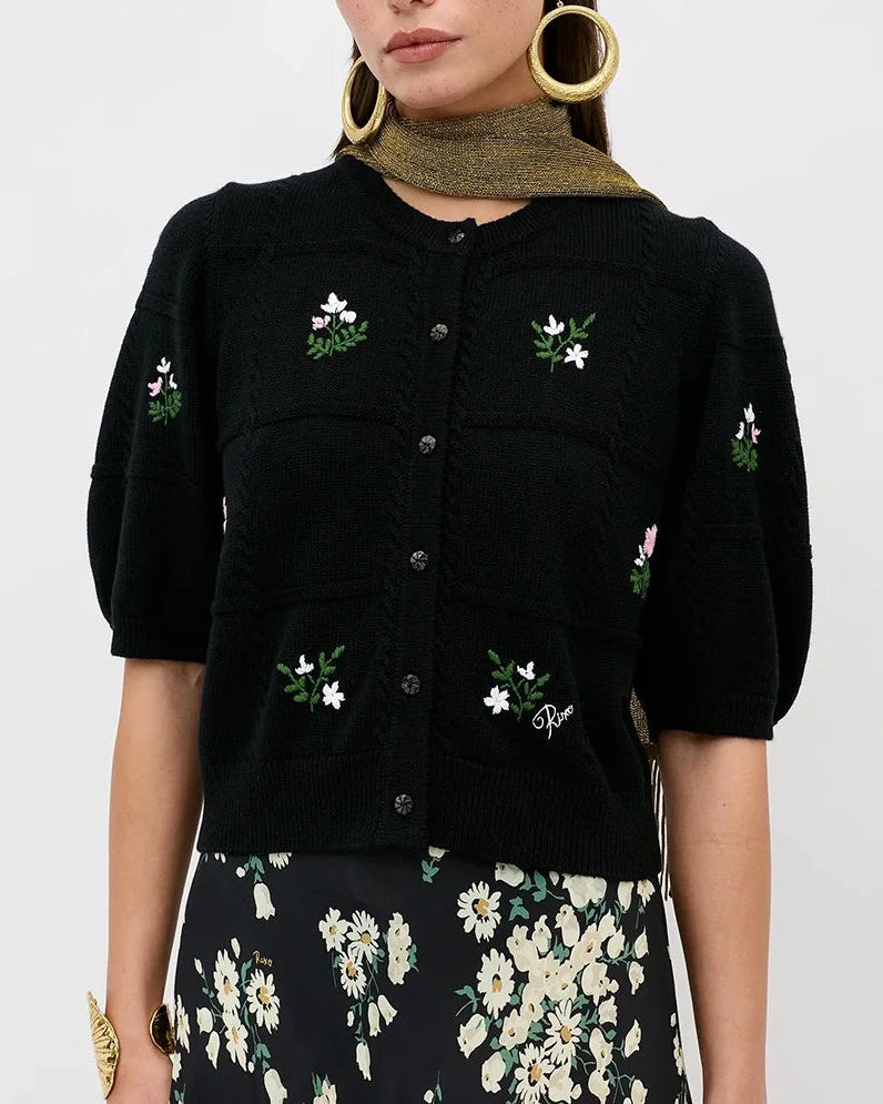 A model wearing a black short-sleeve button-front cardigan with white and green floral embroidery, paired with a black midi skirt featuring a white floral print and a green ribbed turtleneck underneath. She is also wearing large gold hoop earrings and gold bracelets, set against a plain light gray studio background.