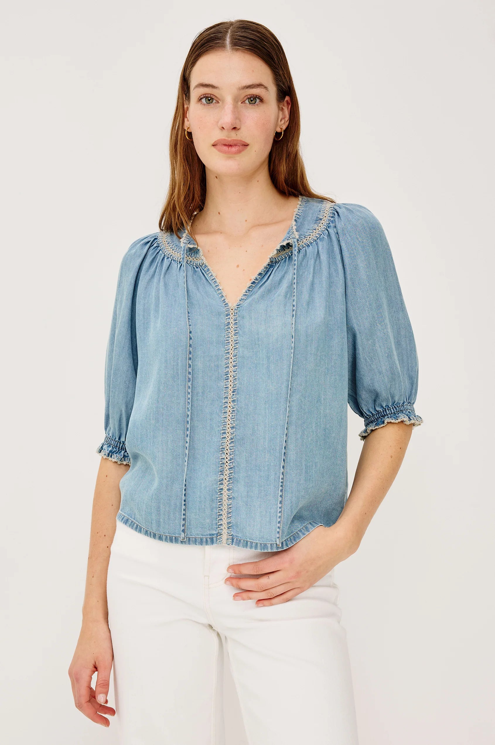 Woman wearing a light blue denim blouse with white pants on a plain background