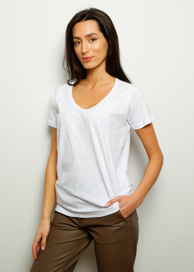 A woman facing away from the camera wears a white v-neck, short-sleeved t-shirt with a relaxed fit. She has dark hair and is wearing brown trousers.